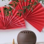 Ceramic incense holder with burning cones, red paper fans, and green leaves on a white background.